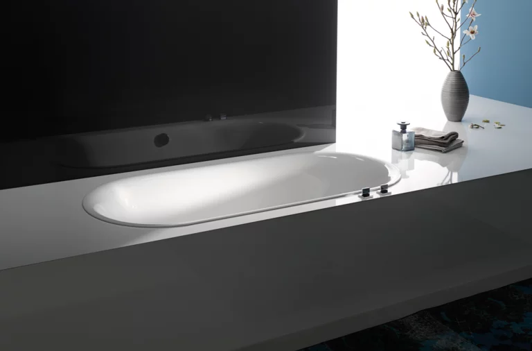 Bañera ovalada de acero esmaltado, integrada a ras en superficie blanca, al lado jarrón con ramas y toallas.
